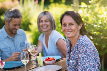 A bunch of friends gathered to share a meal around a table in the garden. Focus on a beautiful woman looking at the camera