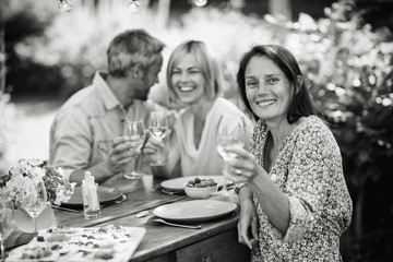 In summer. a group of friends in their forties gathered around a table in the garden to share a meal. They toast with their glasses of wine to the camera.