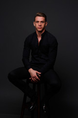 Fototapeta na wymiar a young, white guy sits on a chair in a black shirt and black pants, against a black background and looking at camera. business, model, interview, portrait