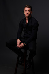 Fototapeta na wymiar a young, white guy sits on a chair in a black shirt and black pants, against a black background and looking at camera. business, model, interview, portrait