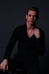 Fototapeta premium a young, white guy sits on a chair in a black shirt and black pants, against a black background,straightens the shirt and looking at camera. business, model, interview, portrait