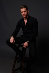 Fototapeta premium a young, white guy sits on a chair in a black shirt and black pants, against a black background and looking at camera. business, model, interview, portrait