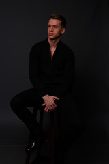 a young, white guy sits on a chair in a black shirt and black pants, against a black background and looking away. business, model, interview, portrait