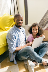 african american father with laptop and daughter with dog sitting on floor together at home