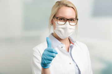 Junge hübsche Ärztin mit Mundschutz und hygienischen Handschuhen zeigt Daumen hoch