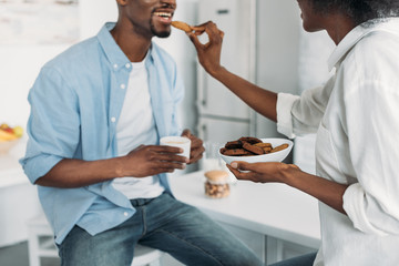 partial view of african american woman feeding husband with cookies in morning in kitchen at home