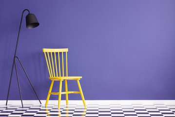 Bright, yellow chair standing next to a black lamp against purple wall in simple room interior. Real photo