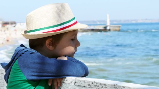Young boy looks thoughtfully at the sea. Side view. Kid in straw hat dreams while walking on a warm spring day near the water