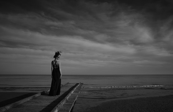 Beautiful Woman In A Dark Luxury Dress With Big Flower Crown On Head Stand On A Wooden Pier. Dark Clouds And Sea On Background. Black And White