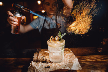 Barman prepares cocktail with orange and herbs in transparent glass on bar with alcohol. Uses burner with sparks. Dark background.