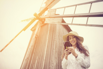 A beautiful young woman is using an application in her smart phone device, in spring time, in front of a windmill.