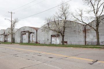 Fototapeta na wymiar A blooming white crabapple trees next to a row of old warehouses. The warehouses are rusted and abandoned, with round roofs. A street runs beside them with a double yellow line and road repair patches