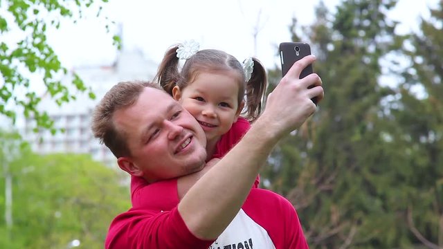 Happy young white family of father and child taking selfie. Dad and daughter embracing and photographing themselves posing for photo outside in green city spring or summer park.