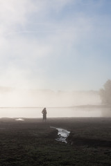 A female silhouette standing near a lake in the morning surrounded by fog, with a stream flowing in the foreground.