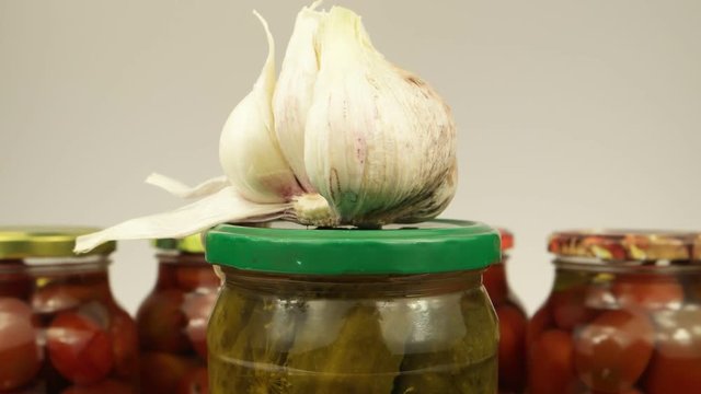 Glass can of canned cucumbers and garlic head in the background of cans with tomatoes