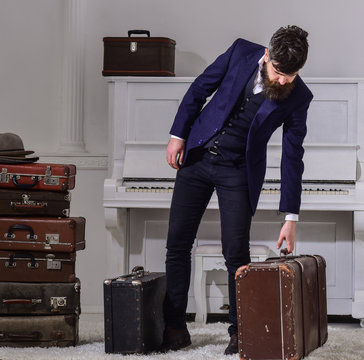 Baggage delivery concept. Man, traveller with beard and mustache with luggage, luxury white interior background. Macho elegant on strict face stands near pile of vintage suitcase, holds suitcase.