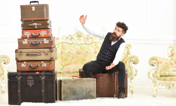 Man, butler with beard and mustache delivers luggage, luxury white interior background. Macho elegant on surprised face sits shocked near pile of vintage suitcase. Luggage and travelling concept.