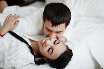 Young husband and wife of white coats play and smile in bed in a hotel room. Close up.