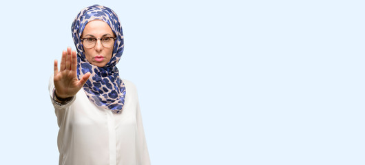 Middle age muslim arab woman wearing hijab annoyed with bad attitude making stop sign with hand, saying no, expressing security, defense or restriction, maybe pushing isolated blue background