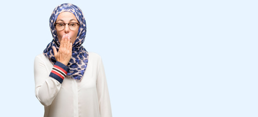 Middle age muslim arab woman wearing hijab covers mouth in shock, looks shy, expressing silence and mistake concepts, scared isolated blue background