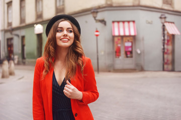 Fototapeta premium Stylish hipster girl posing in front of the old street in city. Trendy young woman model. Street fashion concept, wearing hat and red jacket. Tourist smile, sunny day.Elegant girl with long curly hair