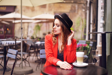 Fototapeta na wymiar Young stylish beautiful woman sitting in city cafe in red jacket, street style, drinking aromatic coffee. Elegant girl in hat smile. Sweet breakfast, happy face, outdoor hipster portrait, fashion girl