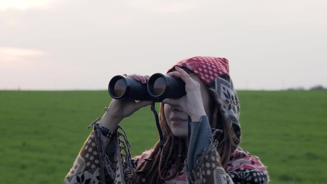 Young readhead woman with dreadlocks  watching in binocular in green summer field against sunset