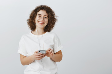 Naklejka premium Sister learns spanish via audiobook. Studio shot of positive good-looking caucasian female student in glasses with short curly hair, holding smartphone, listening songs in earphones over gray wall