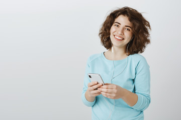 Taking break with music in ears. Shot of pleased carefree european female friend in casual clothes, listening songs in earphones and holding smartphone, staying in touch with family via internet