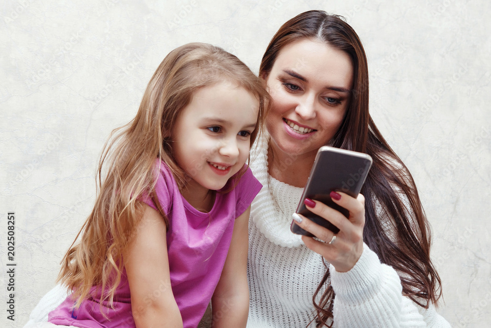 Wall mural young woman and girl are looking together at the mobile phone screen. they are sitting at a table on