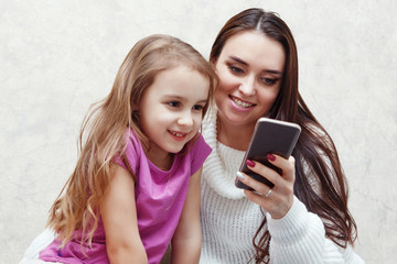 Young woman and girl are looking together at the mobile phone screen. They are sitting at a table on which there is a laptop. It's mom and daughter.