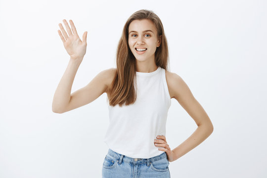 Portrait of confident happy young fair-haired woman in tank-top, holding hand on hip and waving with palm at camera, smiling broadly, saying hi or hello while greeting friend on street over gray wall