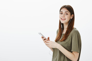 Studio portrait of pleased confident young european woman in dark-green t-shirt, gazing at camera while picking song in smartphone, listening music in earphones over gray background