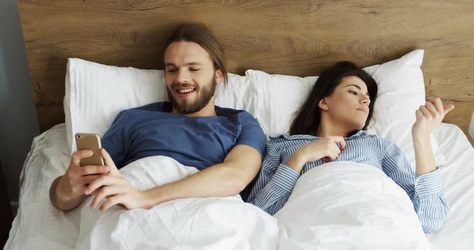 Young caucasian couple lying in the bed in the morning, man using a smartphone, then hugging his wife and making selfies of them. Indoor