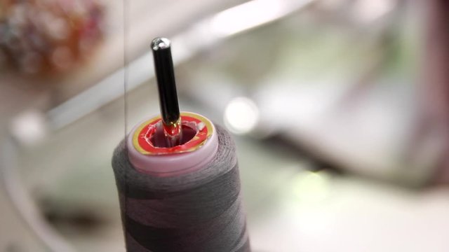 Skilled seamstress work with a grey cloth on professional sewing machine under artificial lightening. A spool of high quality grey cotton thread on a sewing machine spindle on foreground, close-up. 