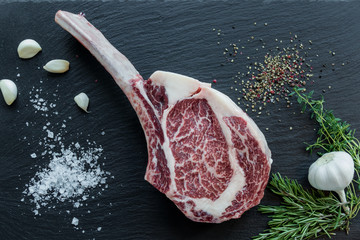 Frisches, rohes Tomahawk Steak mit Kräutern und Gewürzen auf einer schwarzen Schieferplatte