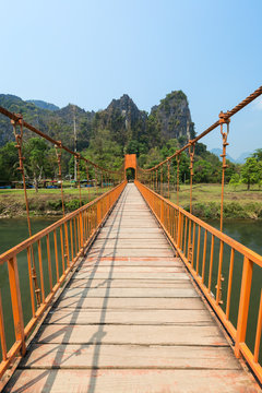 Orange suspension footbridge over the Nam Song River and scenic limestone karst mountains near the Tham Chang (or Jang or Jung) Cave in Vang Vieng, Laos, on a sunny day.