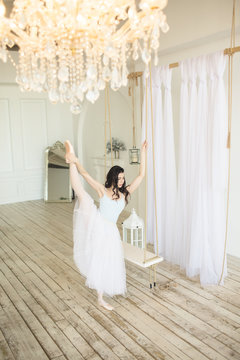 Young pretty ballerina is wearing white skirt dancing near swing in studio with light interior and wooden floor