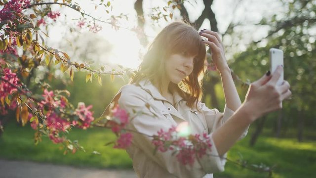 beautiful young red-haired woman among cherry blossom sakura tree spring pink flowers making selfie on smartphone on sunset 