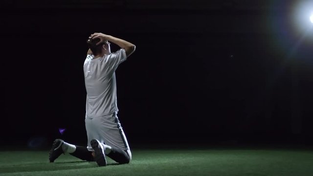 Rear view of soccer player falling on knees on field in dark arena with lightning projector and expressing disappointment about team failure