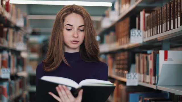 Thoughtful young female student reads a book standing in the library