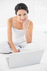 Obraz na płótnie Canvas Cheerful casual brunette in white pajamas sitting on her bed and using a laptop