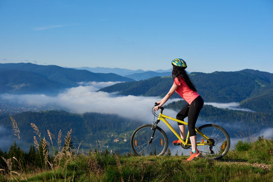 Sporty female cyclist riding on yellow bicycle on the top of mountain, wearing helmet and red red t-shirt, enjoying morning haze in valley, forests on the blurred background. Outdoor sport activity