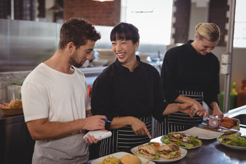 Cheerful chef pointing at food to waiter by female colleague