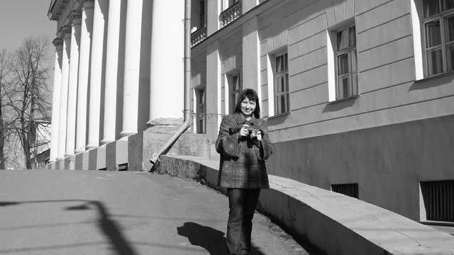 Young woman tourist in vintage gray coat photographing architecture in empire style on retro camera,black and white shooting.