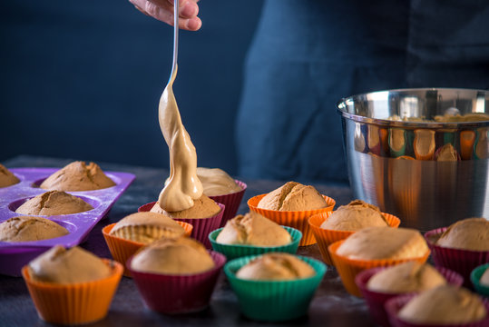 Chef pastry is pouring cream on freshly prepared cupcakes on dark background. Concept of confectionery cooking