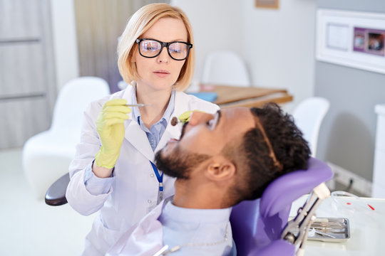 Attractive young dentist wearing white coat and rubber gloves examining oral cavity of mixed-race patient while having appointment at modern office