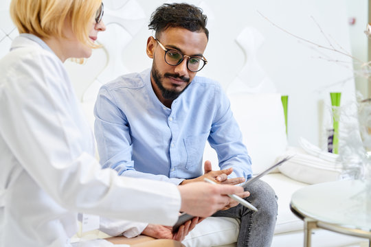 Handsome mixed-race patient answering questions of female physician while she filling in medical record, interior of modern office on background