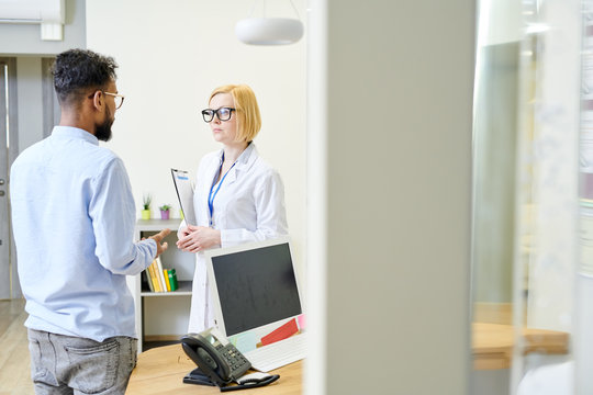 Pretty blond-haired physician wearing white coat and eyeglasses standing at spacious office while consulting bearded patient