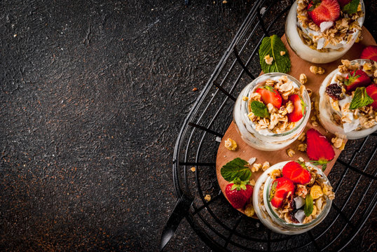 Healthy summer breakfast idea, homemade layered parfe dessert in small jar with granola, yoghurt and strawberry, dark background copy space